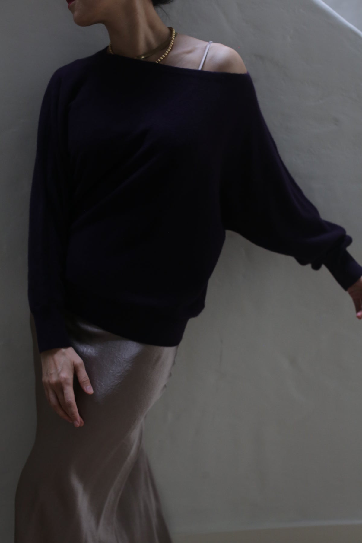 Clad in the Ballerina's Cashmere Sweater in Aubergine and a shimmering skirt, an individual poses against a plain wall. Their right arm is gently extended, and their head is turned, partially out of the frame. This sophisticated ensemble flawlessly merges comfort with elegance.