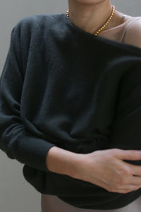 Wearing an olive Ballerina’s Cashmere Sweater with a dark, boat neck design and relaxed fit, a person stands against a neutral background, one arm crossed over their body, accessorized with a gold necklace.