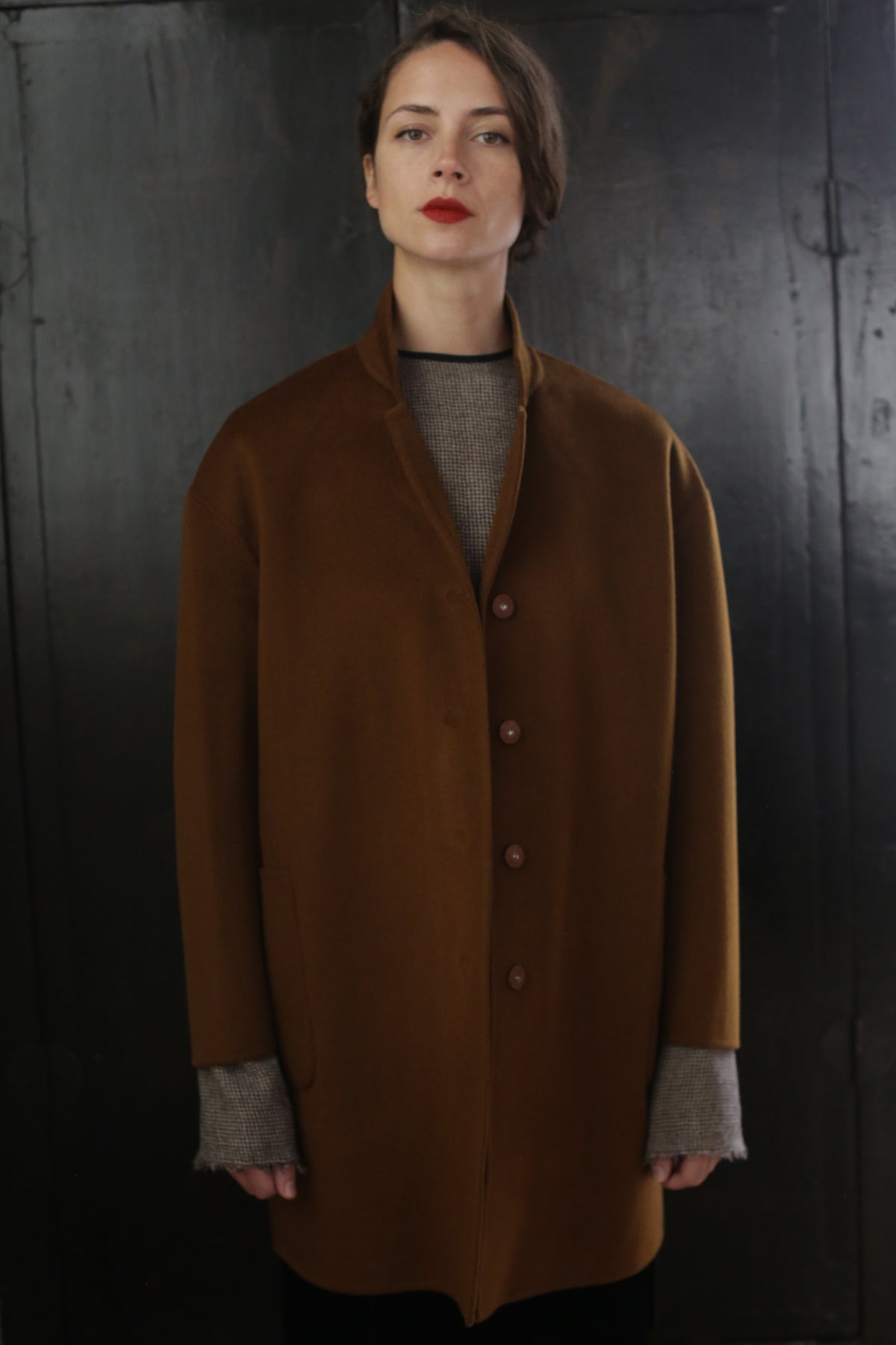 A person with short hair and red lipstick stands against a dark background, wearing a Bamford Double-face Cashmere | Tobacco. The gray sleeves of an inner garment peek out from the coat's sleeves. The person has a calm and composed expression.