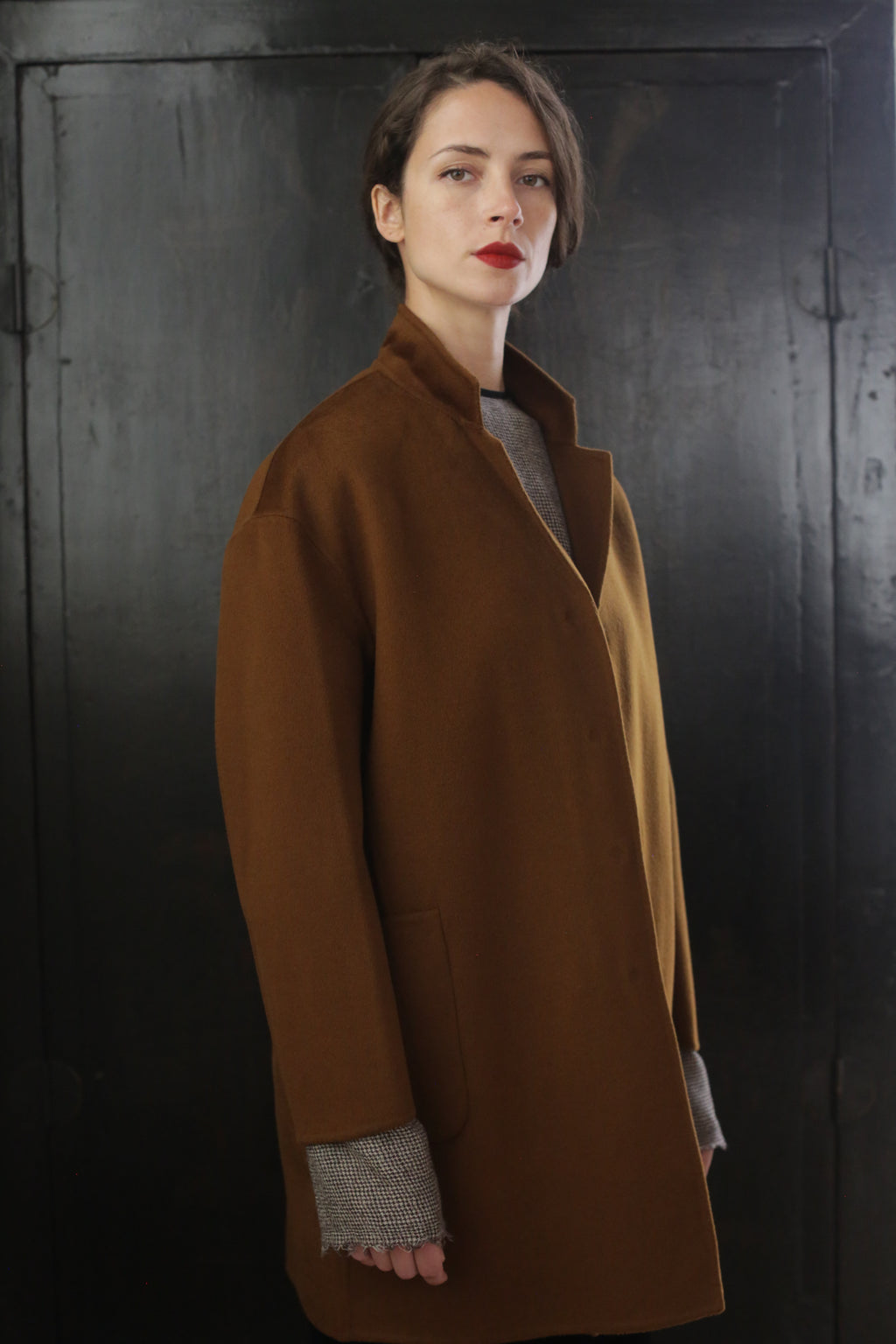 A woman with short dark hair, wearing a Bamford Double-face Cashmere | Tobacco over a gray sweater, stands in front of a dark, textured background. She looks directly at the camera with a neutral expression, and her red lipstick adds a pop of color to her look.