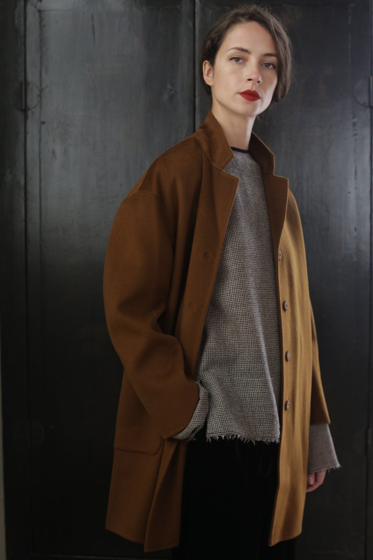 A person with short, dark hair poses in a Bamford Double-face Cashmere | Tobacco over a light-colored, textured sweater. They stand in front of a dark, minimalist background with a neutral expression, looking directly at the camera.
