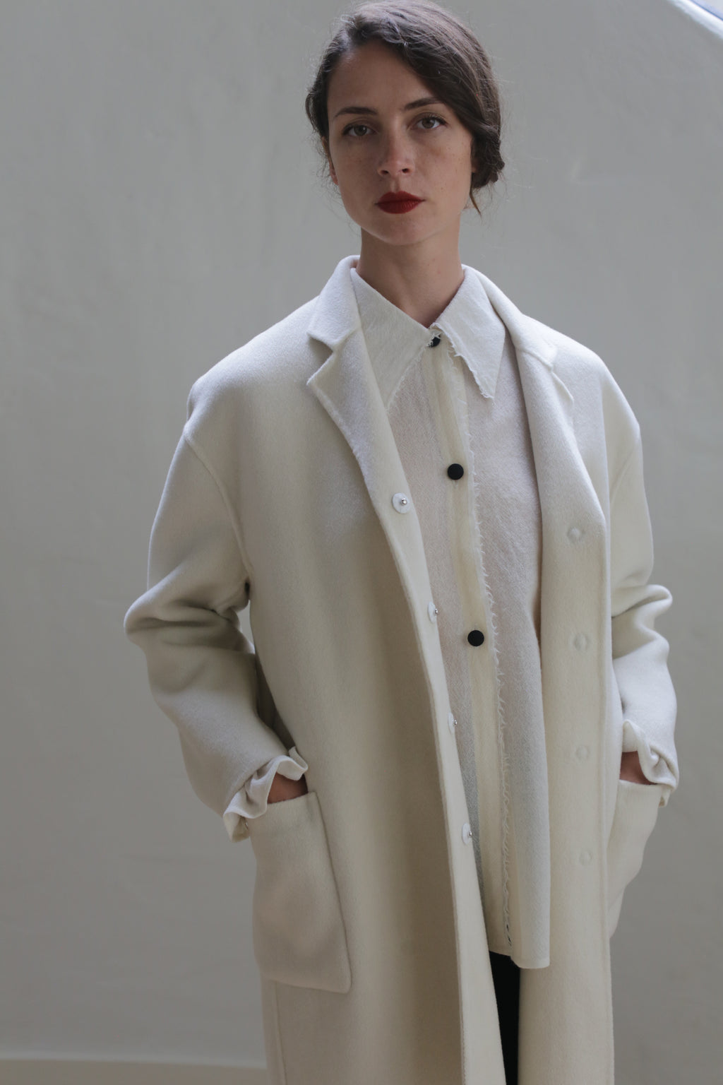 A person stands against a plain backdrop wearing a Bamford Double-face Cashmere | Undyed oversized blazer and a matching button-up shirt. Their hands are in the pockets, they have dark hair pulled back, and they are looking directly at the camera with a calm expression.