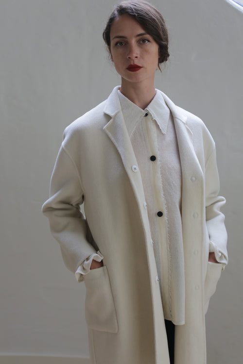 A person stands against a plain backdrop wearing a Bamford Double-face Cashmere | Undyed oversized blazer and a matching button-up shirt. Their hands are in the pockets, they have dark hair pulled back, and they are looking directly at the camera with a calm expression.