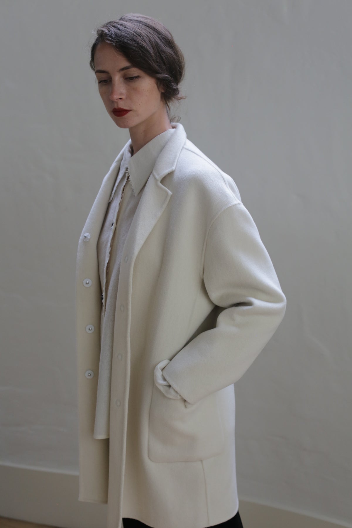 A person with short dark hair is standing against a plain, light-colored background. They are wearing a long, cream-colored Bamford Double-face Cashmere | Undyed coat over a white shirt. One hand is in a pocket of the coat, and they have a composed expression with their gaze directed downward.