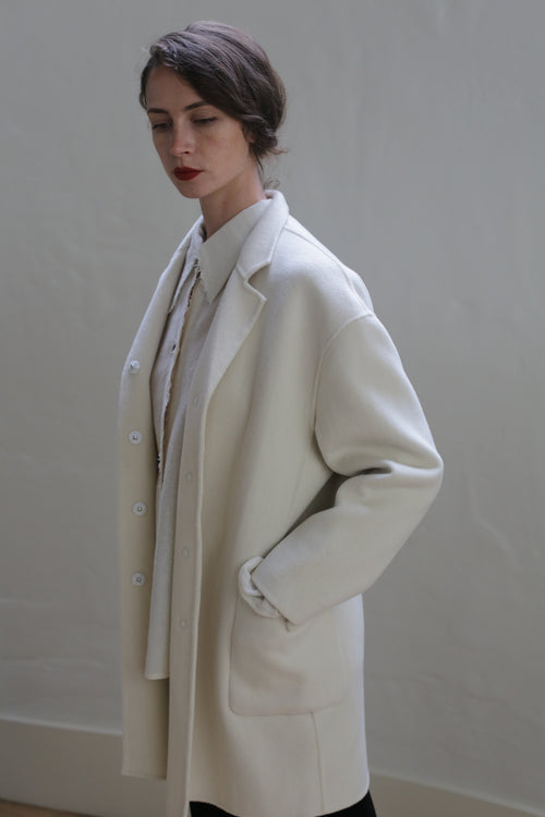 A person with short dark hair is standing against a plain, light-colored background. They are wearing a long, cream-colored Bamford Double-face Cashmere | Undyed coat over a white shirt. One hand is in a pocket of the coat, and they have a composed expression with their gaze directed downward.