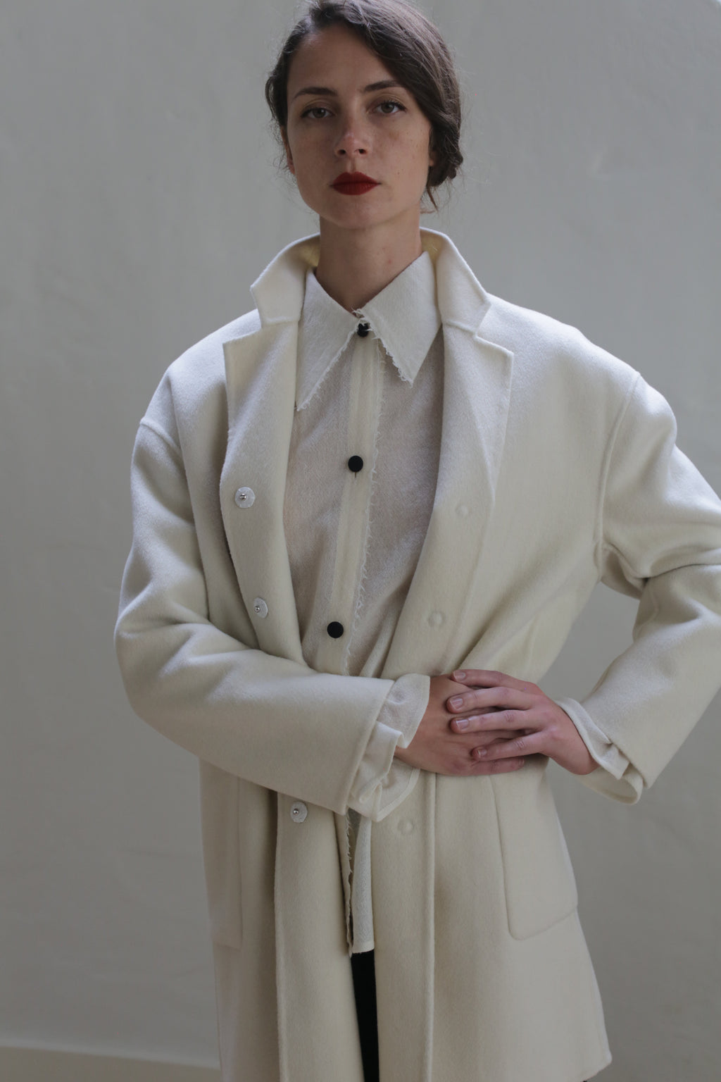 A woman with dark hair, styled in a side bun, stands against a plain background. She is wearing the Bamford Double-face Cashmere | Undyed, layered over a sheer white blouse with a buttoned collar and black buttons. Her hands are clasped at her waist, and she has a serious expression.