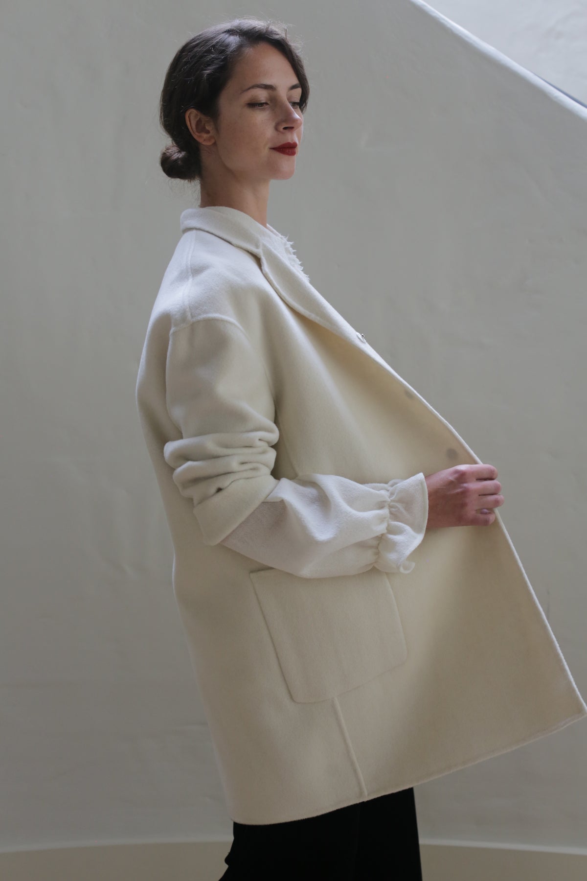 A woman with a serene expression stands against a plain, light background. Her dark hair is pulled back, and she's wearing a loose, off-white Bamford Double-face Cashmere | Undyed coat over a white blouse. With her eyes closed and hands clasping the versatile coat's lapels, she exudes effortless elegance.