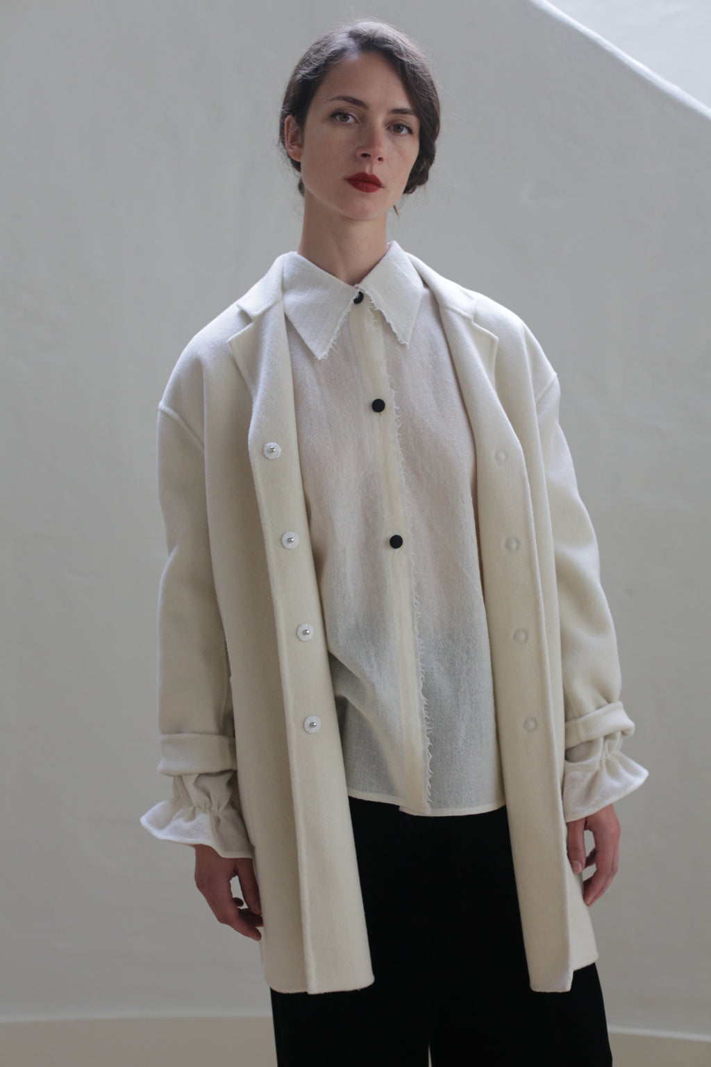 A person with short dark hair stands against a plain light background, adorned in a sheer white blouse with black buttons and an oversized blazer made of Bamford Double-face Cashmere | Undyed. Paired with dark pants and a cream coat with rolled sleeves, their expression is neutral, emphasizing a minimalistic and versatile style.