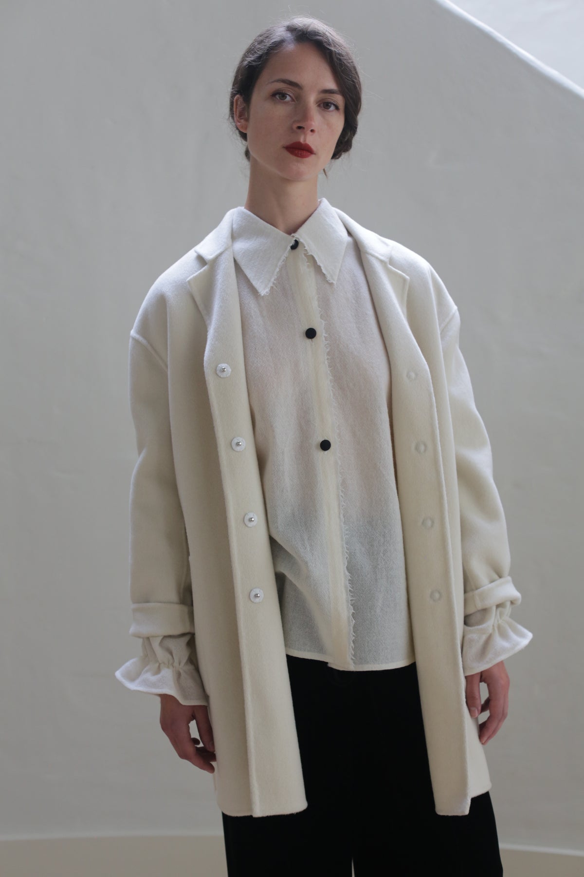 A person with short dark hair stands against a plain light background, adorned in a sheer white blouse with black buttons and an oversized blazer made of Bamford Double-face Cashmere | Undyed. Paired with dark pants and a cream coat with rolled sleeves, their expression is neutral, emphasizing a minimalistic and versatile style.