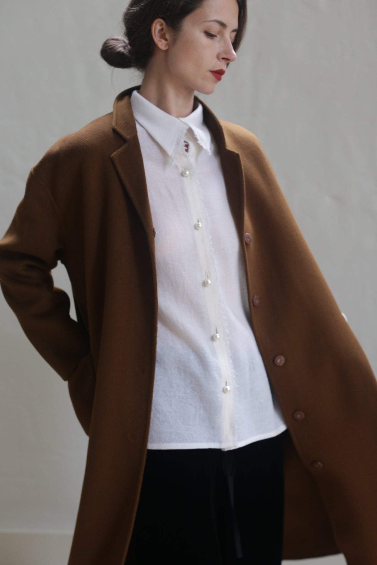 A woman stands with her hands in her pockets, wearing a light brown Bamford Long Double-face Cashmere | Tobacco over a white button-up shirt. Her dark hair is pulled back, and she has red lipstick. The background is plain and neutral, keeping the focus on her outfit crafted from warm material.