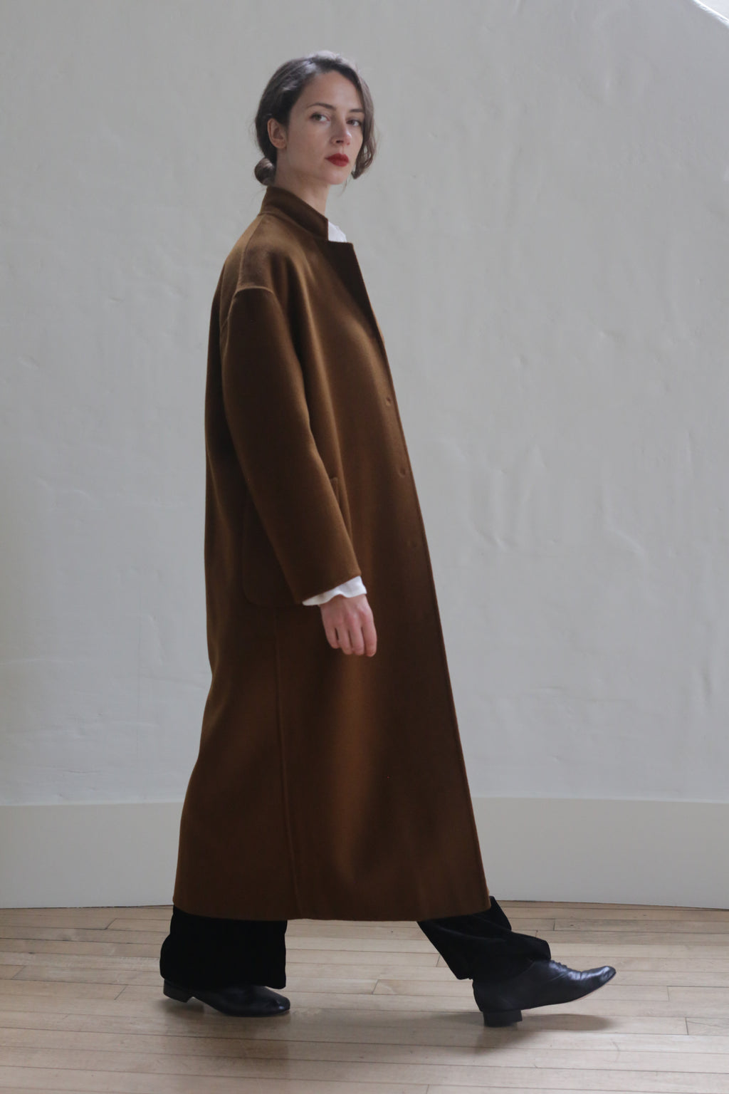 A person wearing a luxurious Bamford Long Double-face Cashmere | Tobacco over a white shirt and black pants stands against a plain white wall. The individual has dark hair pulled back and is looking slightly to the side. Black shoes complete the outfit. They are indoors on a wooden floor.
