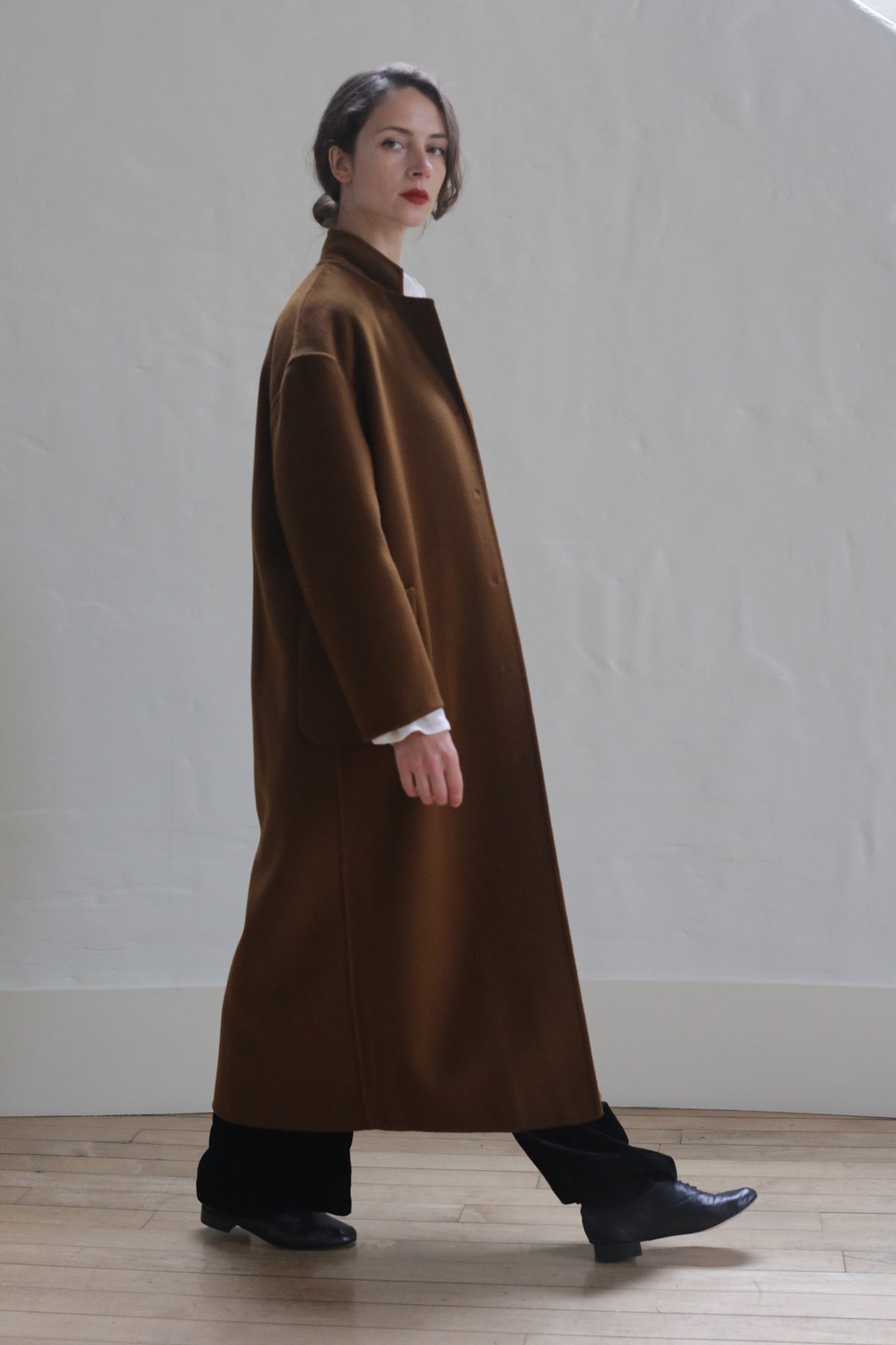 A person wearing a luxurious Bamford Long Double-face Cashmere | Tobacco over a white shirt and black pants stands against a plain white wall. The individual has dark hair pulled back and is looking slightly to the side. Black shoes complete the outfit. They are indoors on a wooden floor.