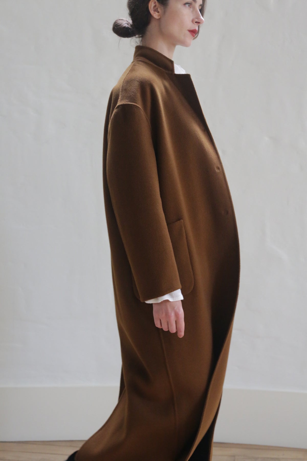 A person with dark hair, styled in a low bun, is wearing a Bamford Long Double-face Cashmere | Tobacco. They stand against a plain, light-colored wall, facing to the side and looking off into the distance. Their hands are in their pockets, and they have a white garment underneath the coat.