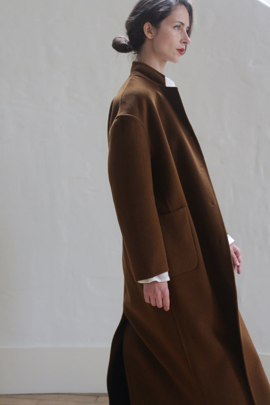 A woman with dark hair tied back is standing sideways, wearing a Bamford Long Double-face Cashmere | Tobacco over a white shirt. She is against a plain, light-colored wall and has a serene expression. The warm material coat features two large pockets and a high collar.