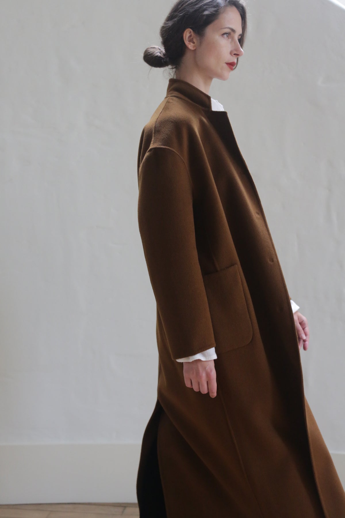 A woman with dark hair tied back is standing sideways, wearing a Bamford Long Double-face Cashmere | Tobacco over a white shirt. She is against a plain, light-colored wall and has a serene expression. The warm material coat features two large pockets and a high collar.