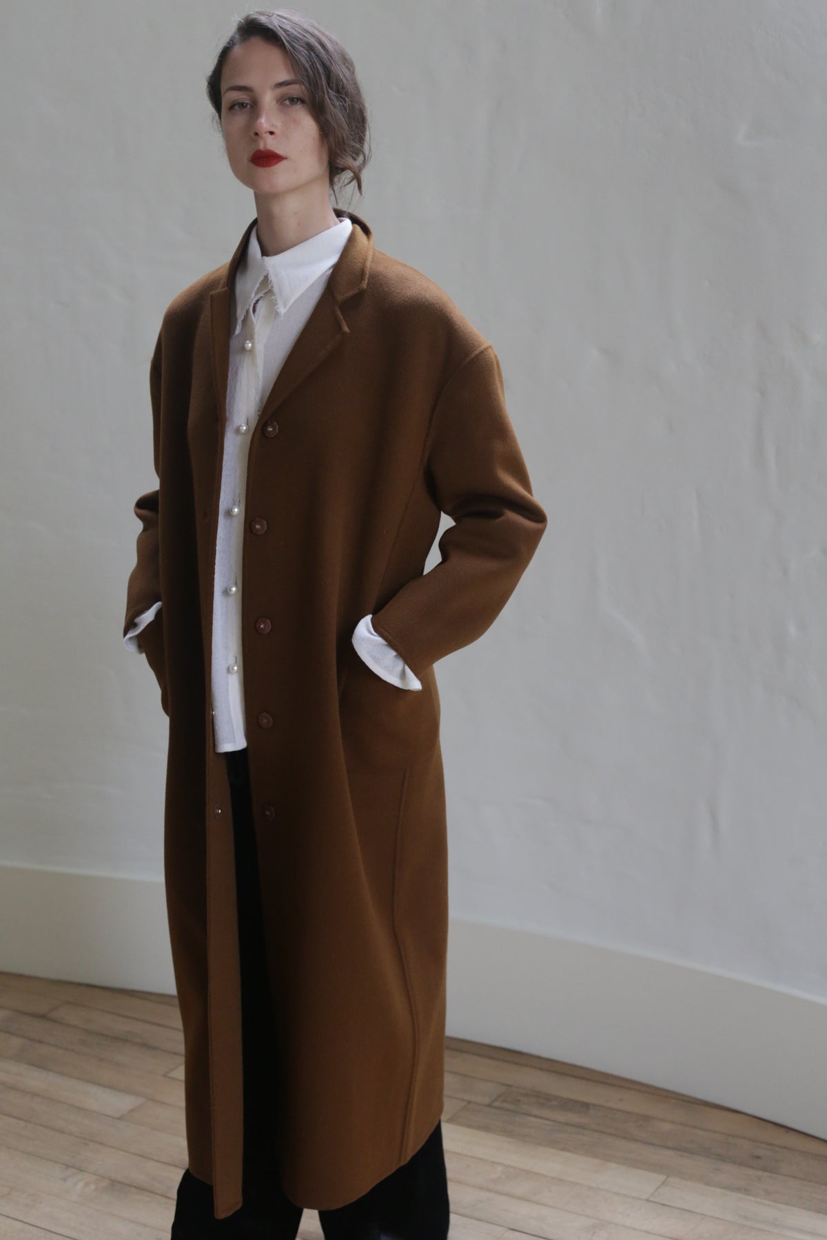 A person with short hair, wearing a Bamford Long Double-face Cashmere | Tobacco coat over a white button-up shirt and black pants, stands indoors against a light-colored wall. They have their hands in their coat pockets and are gazing directly ahead with a neutral expression.