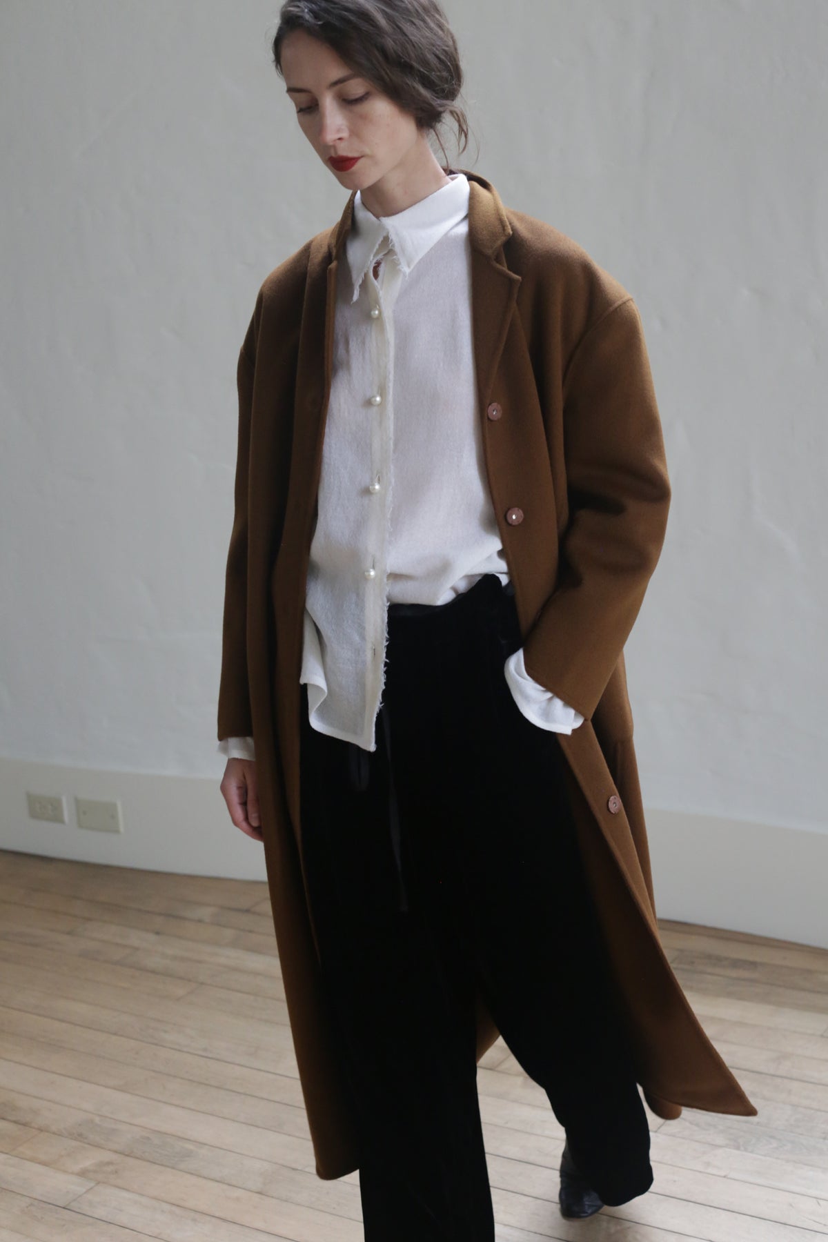 A person with long dark hair is wearing a Bamford Long Double-face Cashmere | Tobacco over a white button-up shirt and black pants. They are standing on a hardwood floor against a plain white wall, looking down and slightly to the side. The setting appears minimalist and modern.