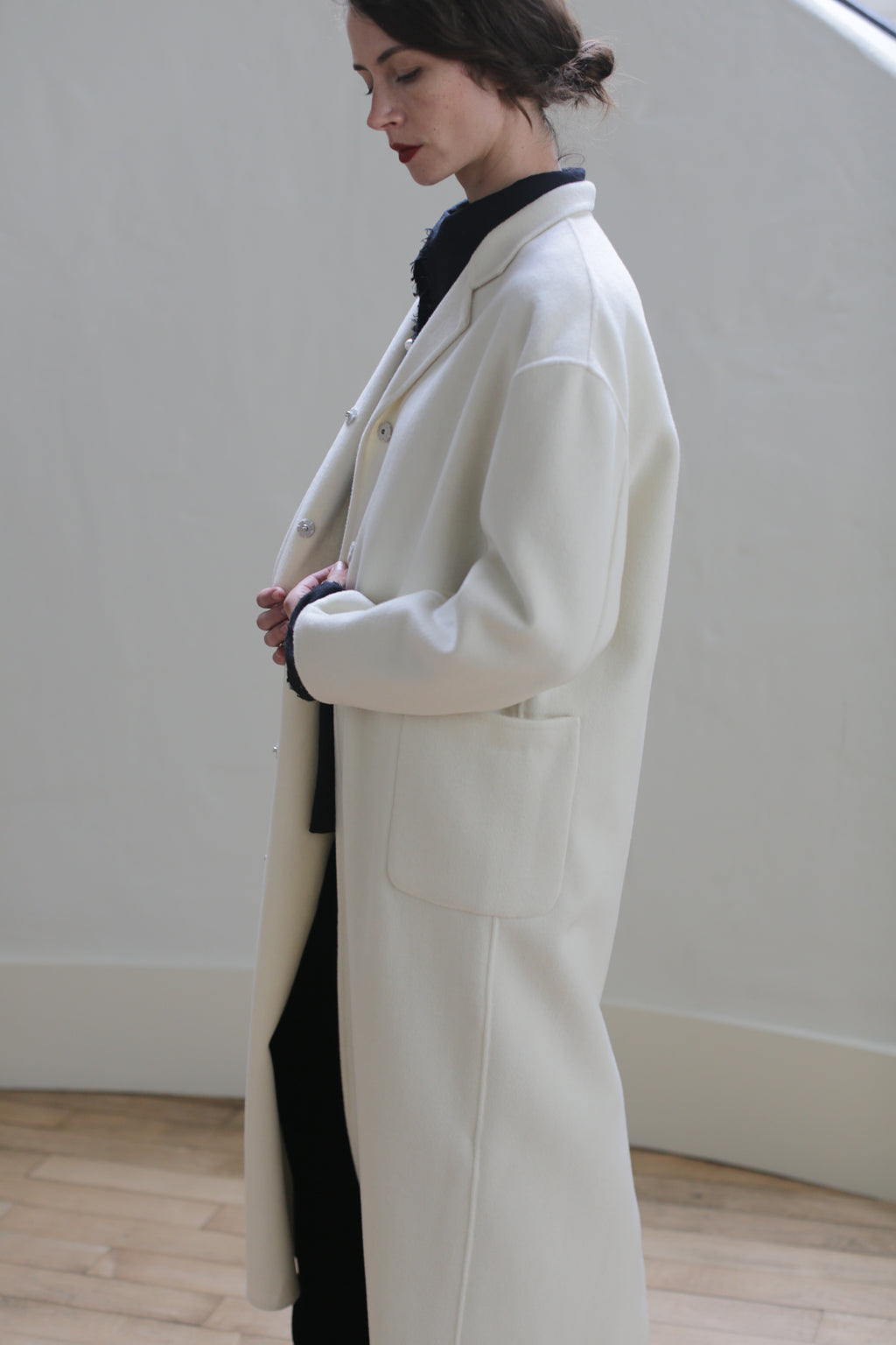 A person with tied-back hair is wearing a Bamford Long Double-face Cashmere | Undyed over a dark outfit. They are standing indoors against a neutral background, facing left, with hands held together at the waist. The setting has minimalistic decor with light wooden flooring and side patch pockets visible.