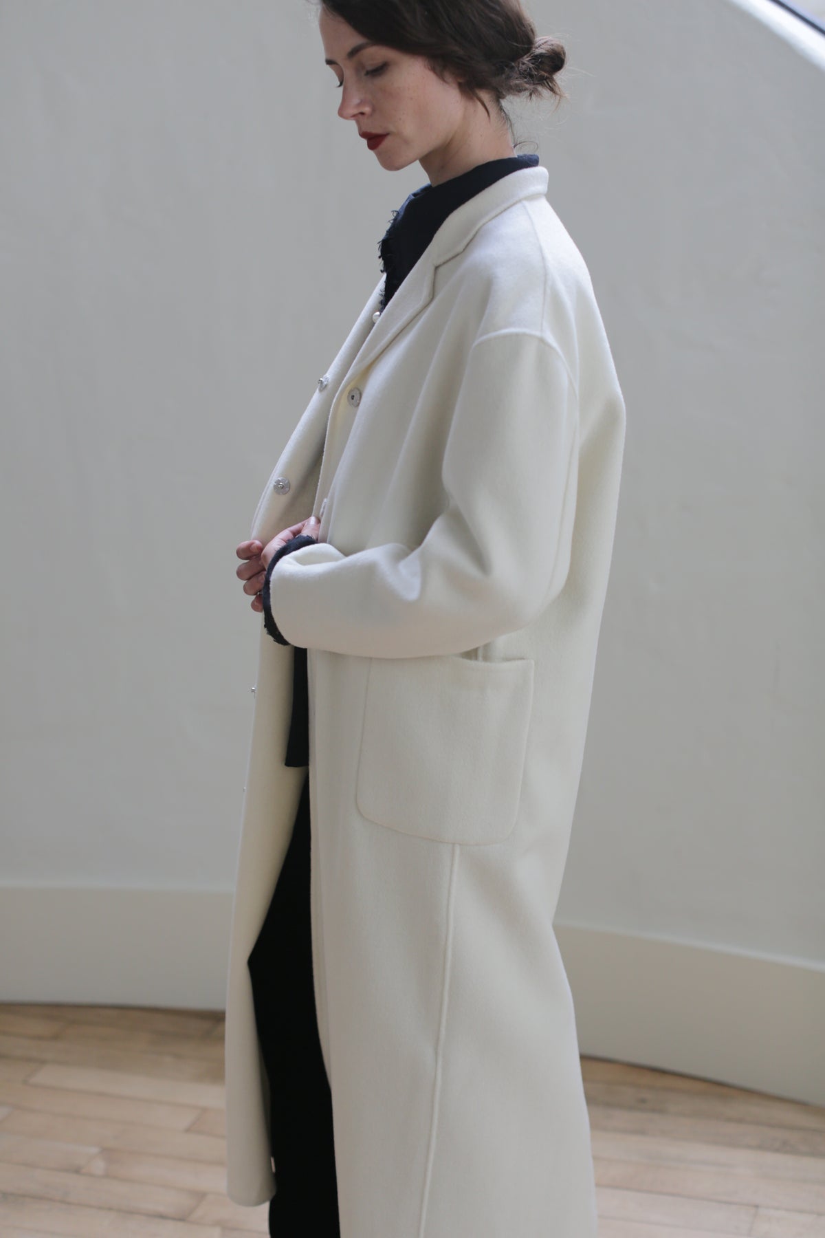 A person with tied-back hair is wearing a Bamford Long Double-face Cashmere | Undyed over a dark outfit. They are standing indoors against a neutral background, facing left, with hands held together at the waist. The setting has minimalistic decor with light wooden flooring and side patch pockets visible.
