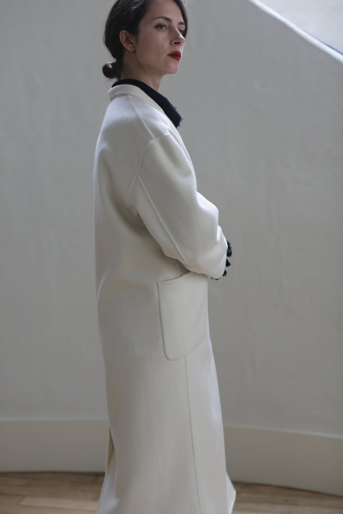 A person stands in profile, dressed in a long, white Bamford Long Double-face Cashmere | Undyed coat with side patch pockets and hands tucked inside. They have short, dark hair and wear a black top underneath. The background is plain, with muted tones, creating a minimalist and elegant setting.