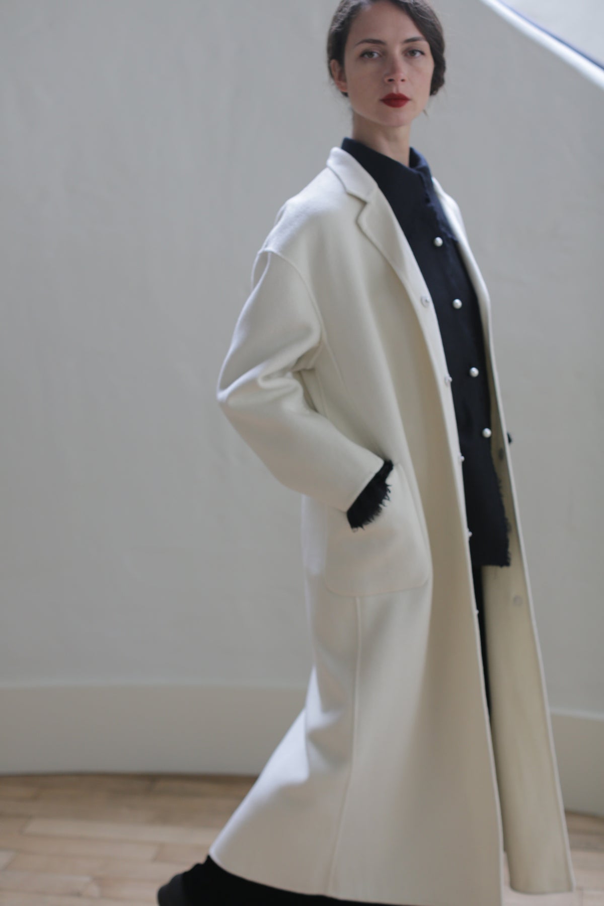 A person with long dark hair and red lipstick is wearing a Bamford Long Double-face Cashmere | Undyed coat with side patch pockets over a dark outfit. They are standing against a plain white wall, looking slightly to the left with one hand in their pocket.