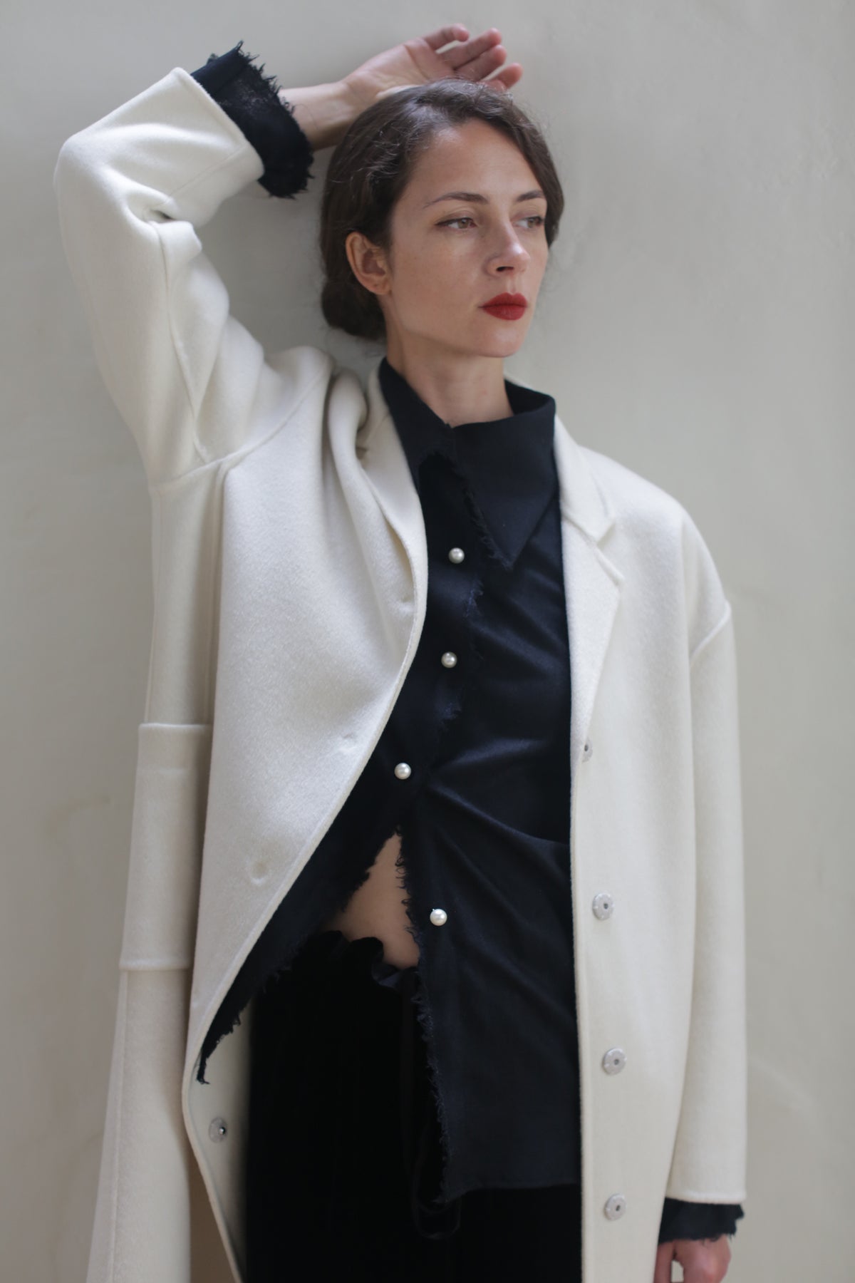 A person stands against a wall with their right arm lifted, resting on their head. They are wearing a Bamford Long Double-face Cashmere | Undyed over a black shirt and have short brown hair, red lipstick, and an attentive expression, gazing to the side. The background is plain and light-colored.