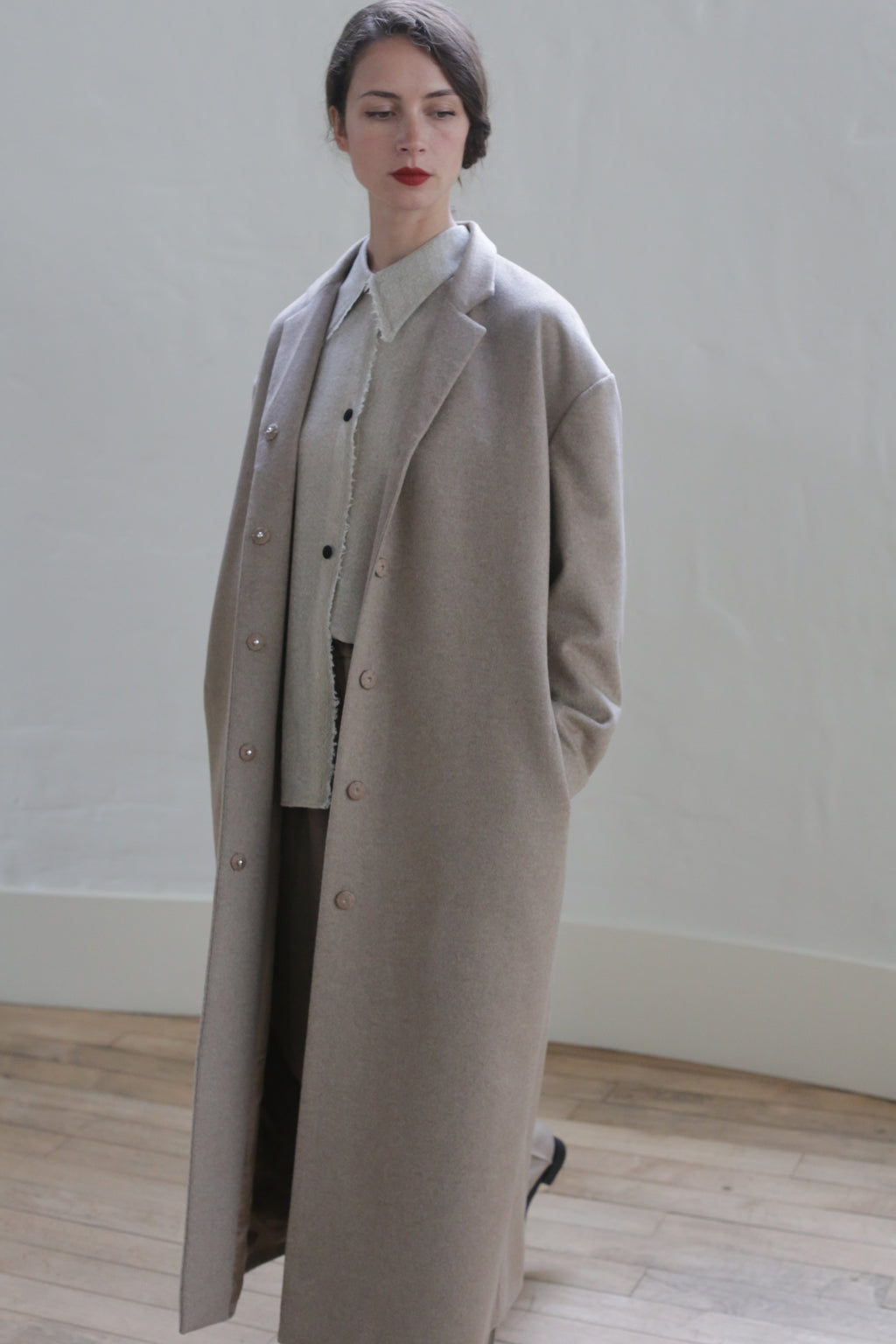 A person with dark hair tied back is wearing a long, light gray overcoat over a shirt and dark pants. They stand in a minimalistic indoor setting with a white wall and wooden floor, hands in pockets, looking off to the side, exuding an air of versatile elegance usually found in the Bamford Long | Undyed Natural.