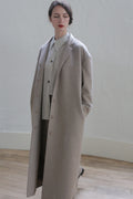 A person with dark hair tied back is wearing a long, light gray overcoat over a shirt and dark pants. They stand in a minimalistic indoor setting with a white wall and wooden floor, hands in pockets, looking off to the side, exuding an air of versatile elegance usually found in the Bamford Long | Undyed Natural.