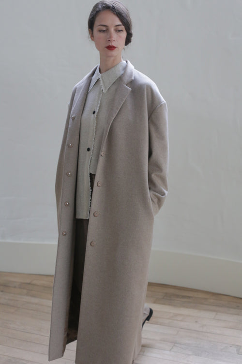 A person with dark hair tied back is wearing a long, light gray overcoat over a shirt and dark pants. They stand in a minimalistic indoor setting with a white wall and wooden floor, hands in pockets, looking off to the side, exuding an air of versatile elegance usually found in the Bamford Long | Undyed Natural.