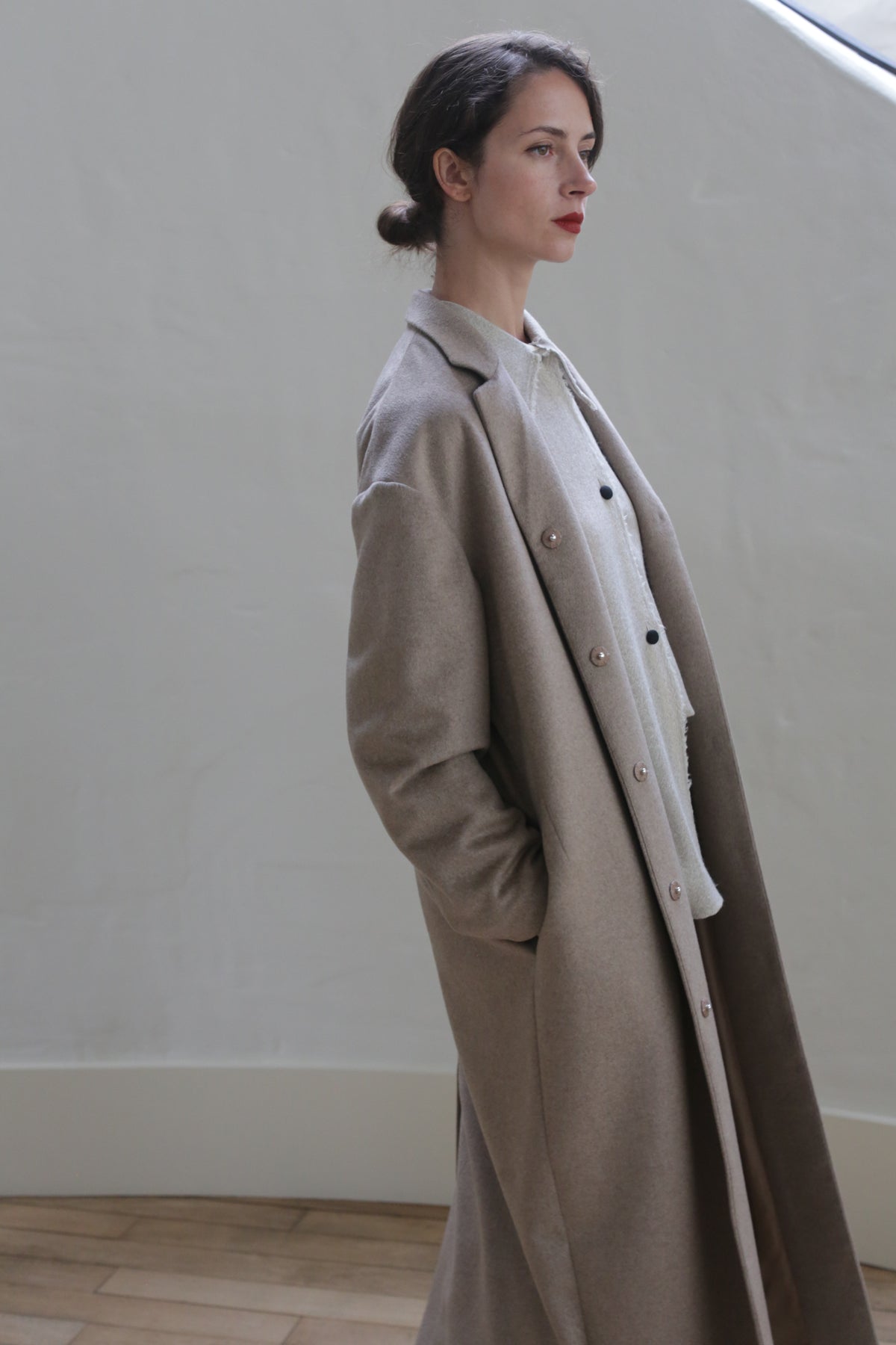 A person with dark hair pulled into a low bun is looking off to the side. They are wearing a Bamford Long | Undyed Natural over a woven cashmere sweater. The background is plain with a soft, neutral tone.