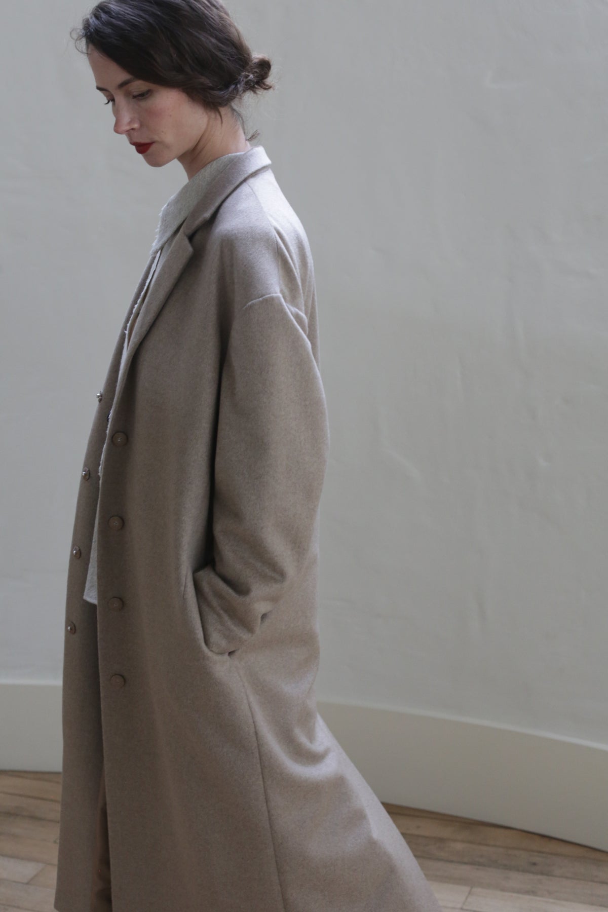 A person with dark hair is standing in a neutral-toned room, wearing the Bamford Long | Undyed Natural with hands in the pockets. The person looks down and away from the camera, creating a contemplative mood. The wall and floor in the background are minimally textured.