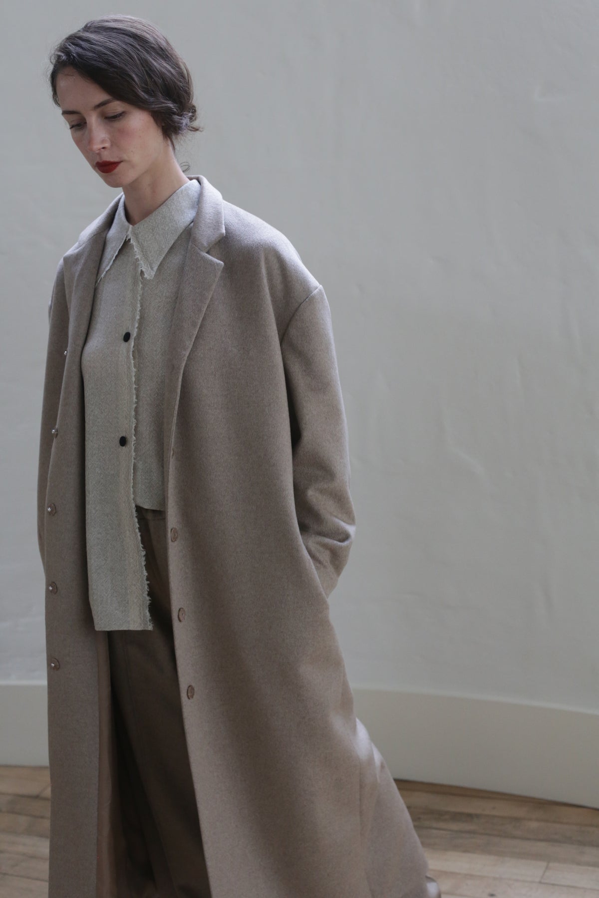 A person with short brown hair stands indoors against a light-colored wall. They are wearing a long, beige coat over a buttoned, collared shirt and wide-legged trousers. Their hands are in their coat pockets, and they are looking slightly downward. The versatile look is enhanced by an oversized blazer made of Bamford Long | Undyed Natural.