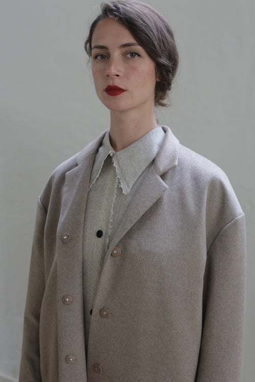 A person with short, dark hair, wearing red lipstick, and dressed in a Bamford Long | Undyed Natural underneath an oversized beige blazer. The background is a plain, light-colored wall. The individual gazes directly at the camera.