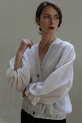 A person with dark hair tied back stands against a plain backdrop, wearing a sheer white blouse with large sleeves and dark buttons. They have a contemplative expression and gaze slightly to the side, with one hand raised and the other resting on their hip, showcasing their handmade cardigan made from Blanc de Chine - Japanese Wool Gauze.