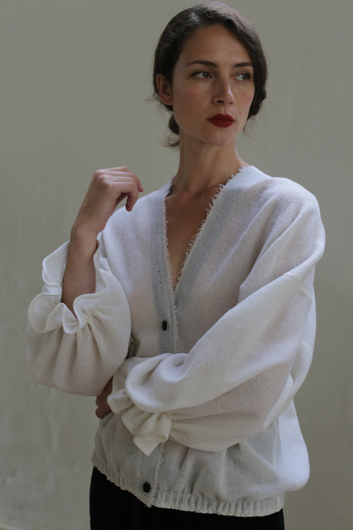 A person with dark hair tied back stands against a plain backdrop, wearing a sheer white blouse with large sleeves and dark buttons. They have a contemplative expression and gaze slightly to the side, with one hand raised and the other resting on their hip, showcasing their handmade cardigan made from Blanc de Chine - Japanese Wool Gauze.