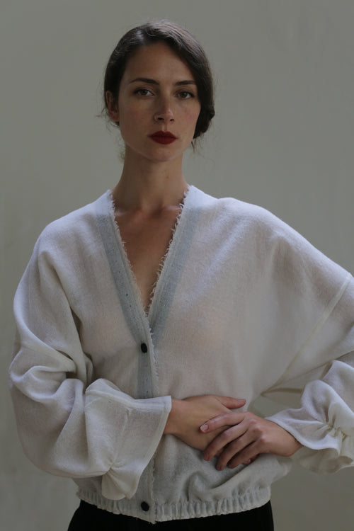 A woman stands with her hands gently clasped in front of her. She has dark hair styled in a loose updo and wears a Blanc de Chine - Japanese Wool Gauze cardigan, featuring light blue trim, large sleeves, and button detailing. She has a neutral expression and stands against a plain background.
