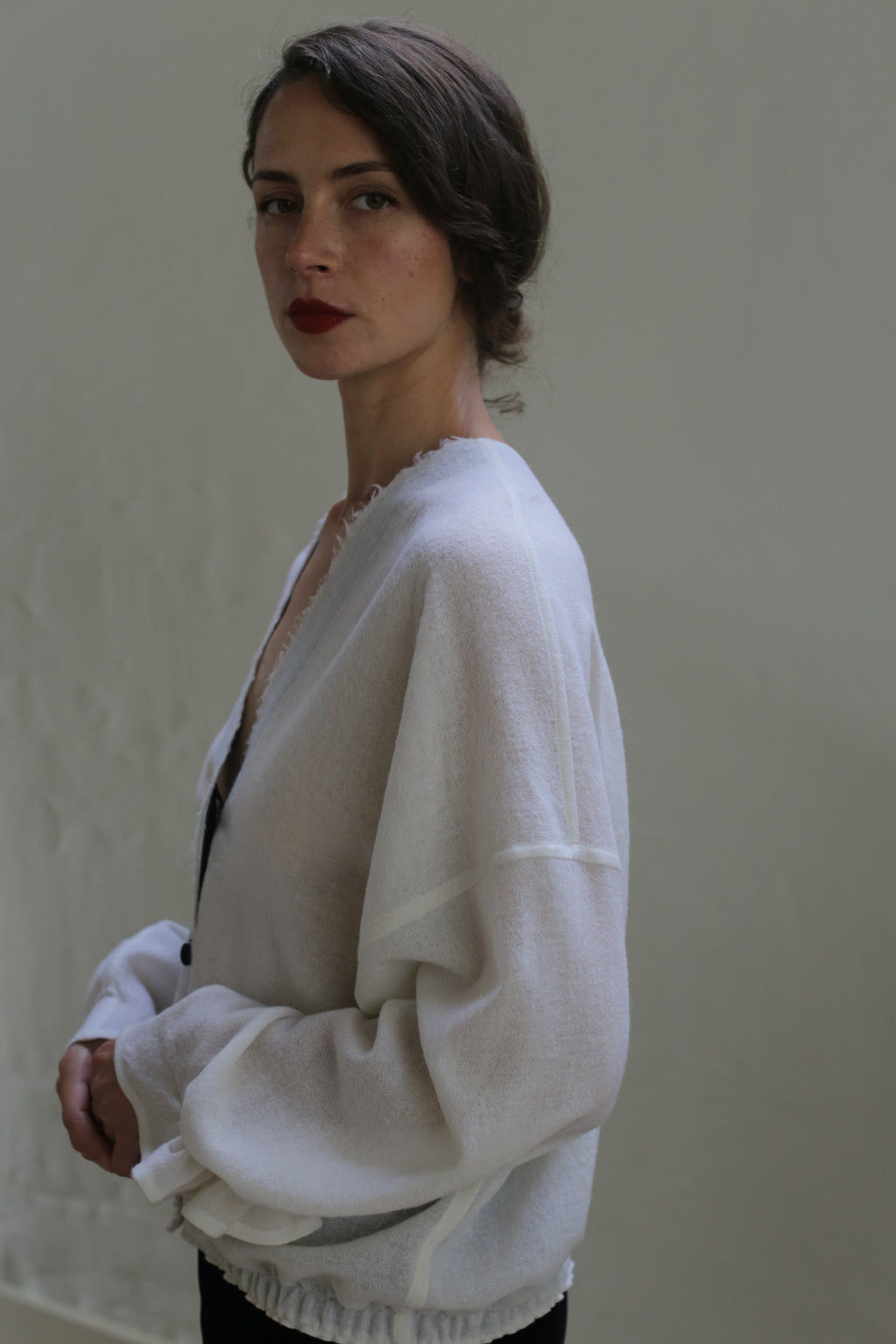 A person with dark hair styled in a low bun is wearing a loose-fitting, handmade cardigan made of Blanc de Chine - Japanese Wool Gauze over a dark top. They are standing against a plain, light-colored background and looking into the camera with a neutral expression.