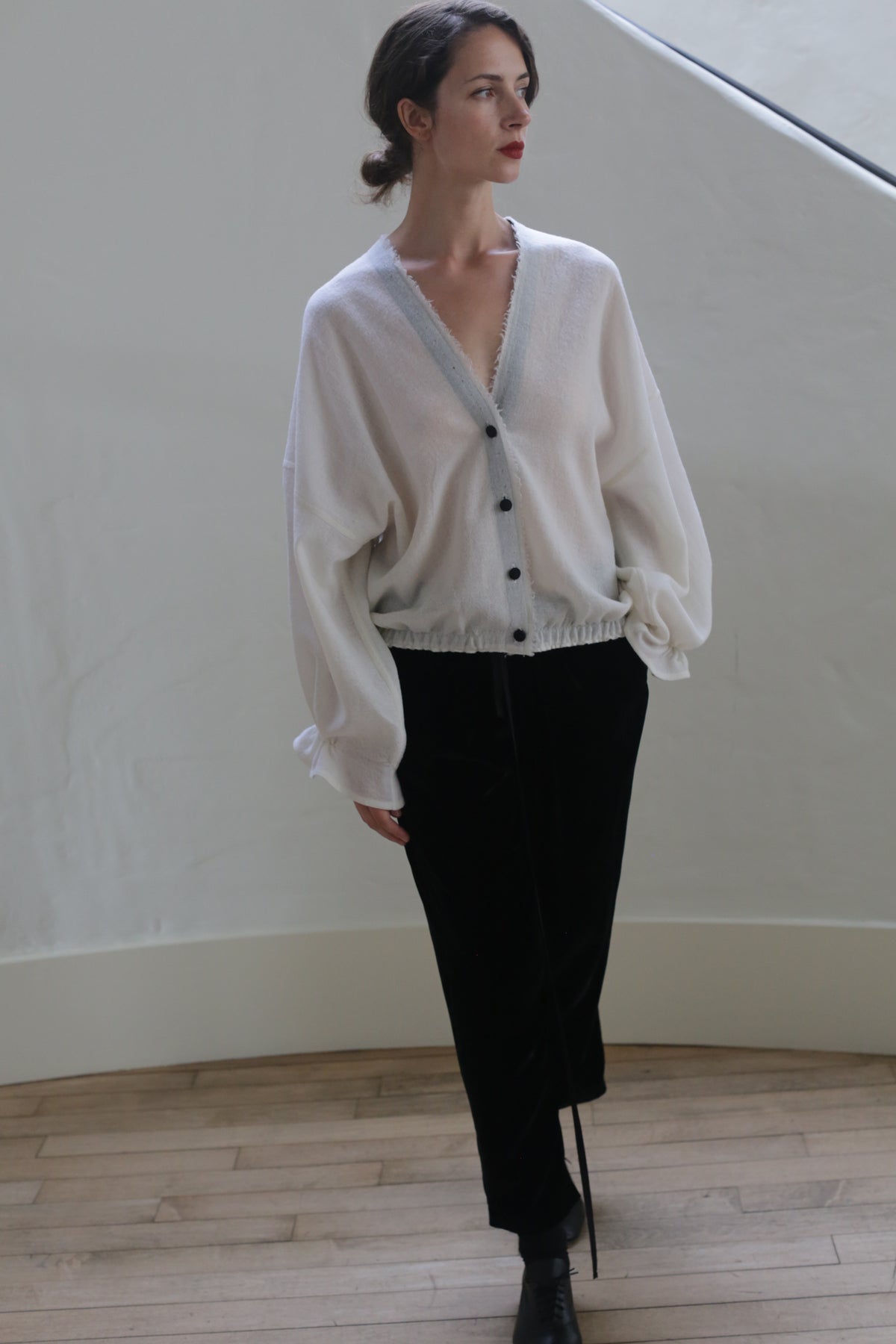 A person wearing a handmade cardigan of Blanc de Chine - Japanese Wool Gauze with black buttons and black pants stands indoors against a light-colored, curved wall. The hair is styled back, and the person gazes to the side with a relaxed posture. The floor is wooden.