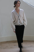 A person with red lipstick and tied-back hair wears a loose-fitting white cardigan with black buttons and Silk Velvet Unisex Pants. They stand on a light wooden floor against a plain, off-white, curved wall, gazing slightly to the side.