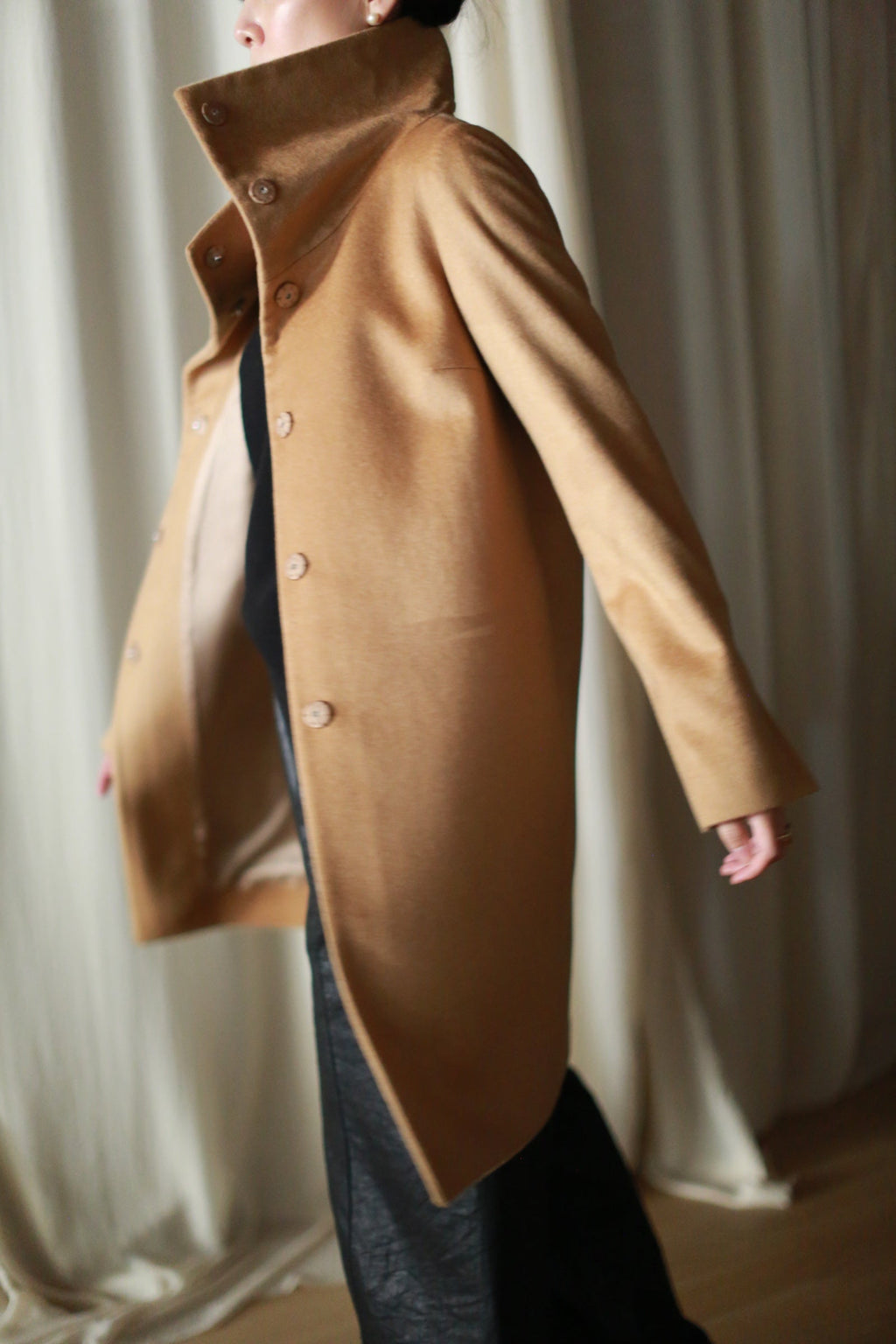 A person wearing a camel-colored Stamford Long | Camel cashmere coat with a straight-cut silhouette, high collar, and front buttons over a black outfit.
