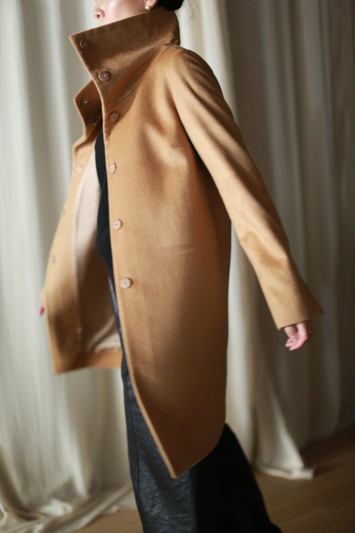 A person wearing a camel-colored Stamford Long | Camel cashmere coat with a straight-cut silhouette, high collar, and front buttons over a black outfit.