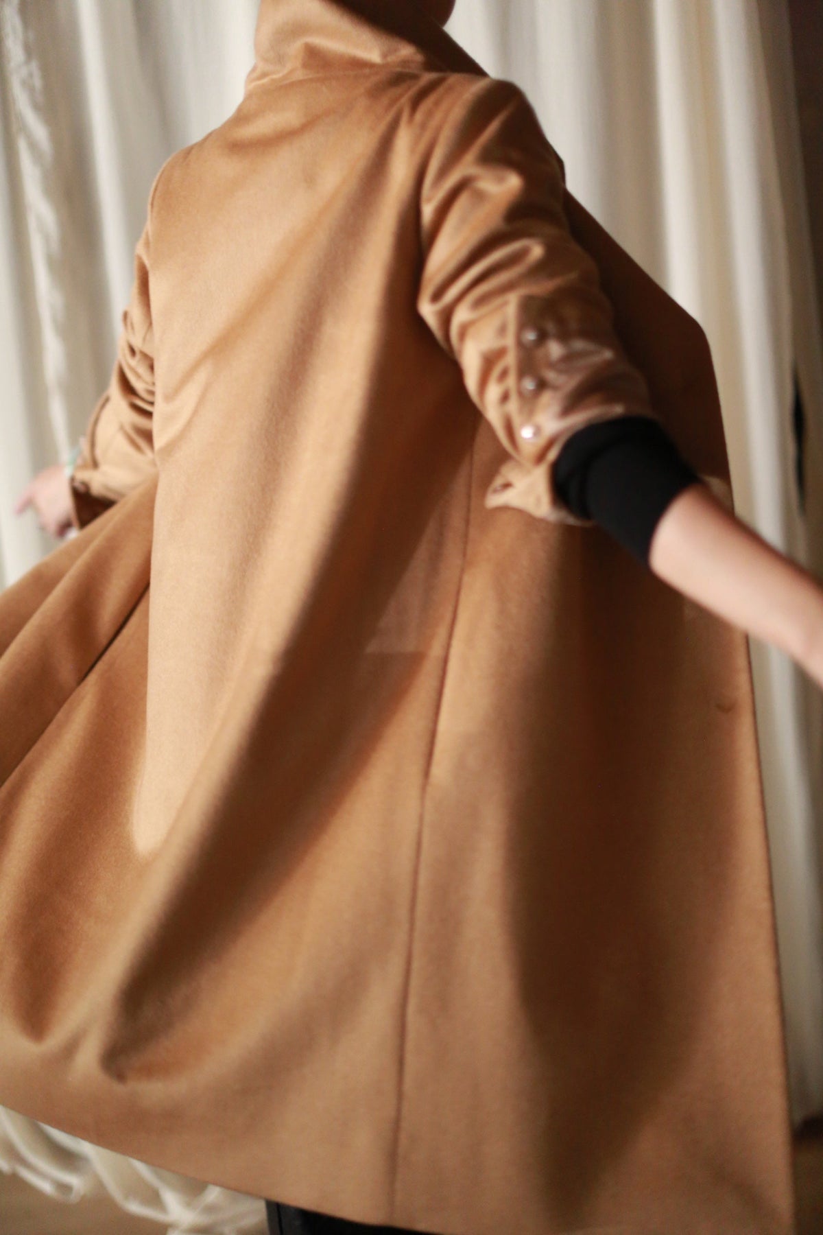 A person viewed from behind wears a straight-cut Stamford Long | Camel coat with a high collar and sleeves rolled up to reveal a black base layer.