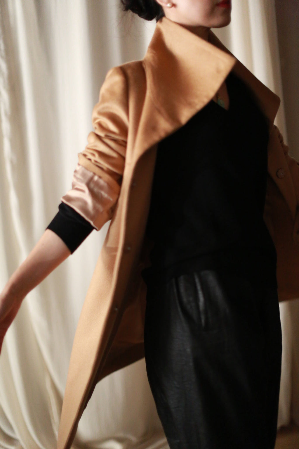 A person wearing a straight-cut, camel-colored Stamford Long coat over a black top and dark trousers.