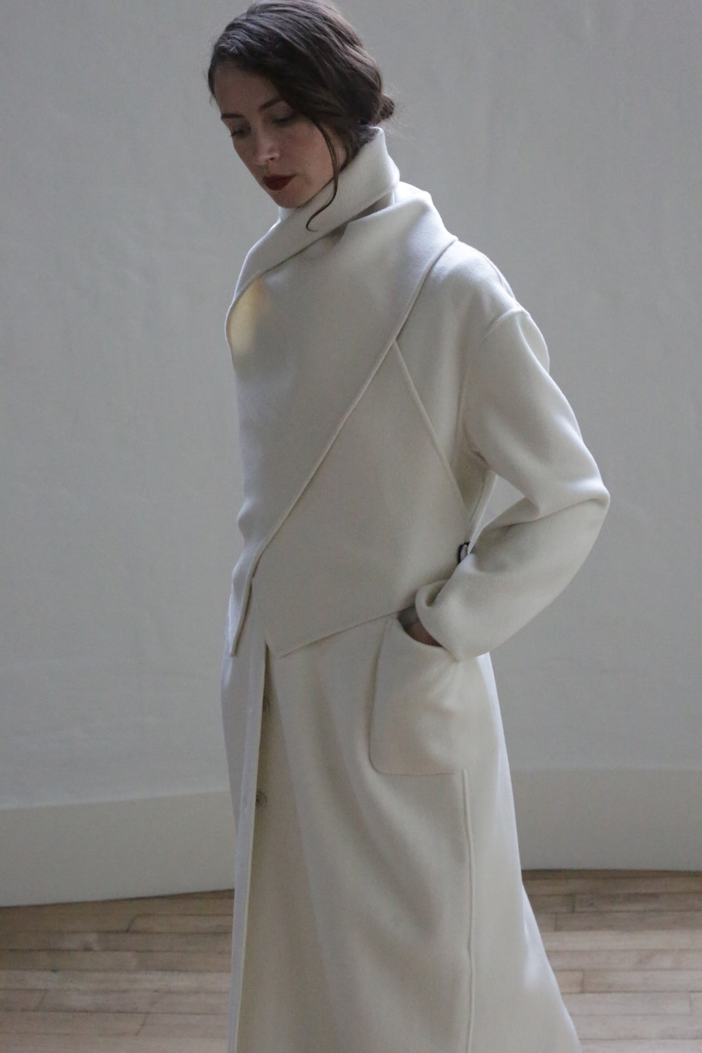 A person wearing a stylish, asymmetrical white coat with ceramic buttons and an X Scarf - Double-face Cashmere | Undyed stands against a plain wall with a wooden floor. The coat has large front pockets, and the individual has their hands in the pockets, looking down slightly. Their hair is tied back, and they have a calm expression.