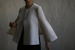 A person wearing the Capelet Long top made from textured, undyed Japanese wool gauze in off-white. The top features flared long sleeves and a round neckline. The background is blurred, revealing a neutral-toned wall. This custom item is accessorized with a gold necklace and bracelets, while their face remains out of view.