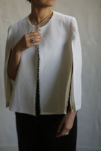 Load image into Gallery viewer, A woman is elegantly dressed in a Capelet Long made from undyed Japanese wool gauze, featuring slit sleeves and black trim. She accessorizes with a beaded necklace and wears a ring on her left hand. The custom-fitted piece drapes perfectly as she grasps the edge of the capelet with her right hand against a plain, neutral background.