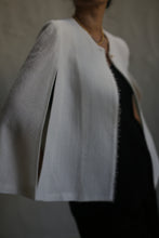 Load image into Gallery viewer, A person is wearing the Capelet Long in Japanese Wool Gauze Undyed, a light-colored, cape-style jacket, over a dark outfit. The custom item features a high collar and open slits along the sides. The background is neutral and out of focus. The person&#39;s face is not visible.