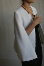 Load image into Gallery viewer, A person is shown from the shoulders down, wearing the Capelet Long made from Japanese Wool Gauze Undyed over a black outfit. They hold the front of the capelet with one hand, revealing a glimpse of a black bra strap. A gold necklace with a pendant is visible around their neck.