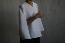 Load image into Gallery viewer, A person wearing the Capelet Long in Japanese Wool Gauze Undyed, along with a necklace, stands against a dark, blurred background. The person&#39;s face is not visible, and their right hand is slightly raised, adorned with rings.