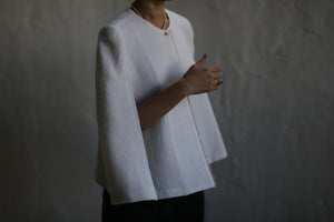 A person wearing the Capelet Long in Japanese Wool Gauze Undyed, along with a necklace, stands against a dark, blurred background. The person's face is not visible, and their right hand is slightly raised, adorned with rings.